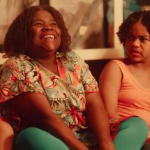 Cena de um dos curtas exibidos no Cineclube Curta o Curta. Duas mulheres negras estão sentadas lado a lado em um ambiente interno iluminado por luz quente. À esquerda, uma delas sorri amplamente e olha para cima, transmitindo alegria. À direita, a outra volta o rosto para a companheira com expressão de surpresa e estranhamento. Ao fundo, uma janela desfocada compõe o cenário.