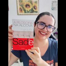 A pesquisadora Nicole Ayres sorri para a câmera enquanto segura o livro A Filosofia na Alcova, do Marquês de Sade, com a capa voltada para a frente. Ela usa óculos de armação escura e blusa azul. O livro, em tons de branco, cinza e vermelho, ocupa boa parte da imagem e destaca o título e o nome do autor.