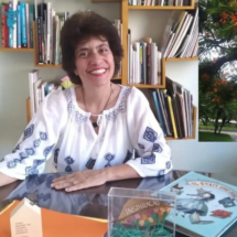 A escritora Marcia Cristina Silva sorri para a câmera enquanto está sentada atrás de uma mesa. Ao fundo, estantes com livros ocupam a parede. Sobre a mesa, há livros infantis, um porta-cartões e uma caixa transparente com a palavra "Inspiração". No canto superior direito da imagem, uma fotografia mostra uma árvore de flores vermelhas à beira de um caminho.