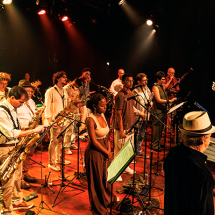 Integrantes da Orquestra Mil Tons se apresentam em um palco iluminado por refletores. O grupo reúne músicos, cantores e instrumentistas distribuídos entre microfones e estantes de partitura. À direita, o maestro rege a apresentação com os braços erguidos, enquanto, ao fundo, diferentes instrumentos de sopro, cordas e percussão compõem a cena. A iluminação em tons quentes reforça o clima vibrante do espetáculo.