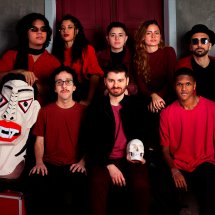 Integrantes da banda autoral Magnífica Máquina Maldita posam em dois níveis diante de uma porta vermelha. O grupo reúne oito jovens com roupas em tons de vermelho, vinho e preto. Em primeiro plano, um dos integrantes segura uma caveira cenográfica, enquanto, à esquerda, uma grande máscara estilizada com traços geométricos completa a composição. Alguns integrantes usam óculos escuros, reforçando a estética contemporânea e teatral da imagem.