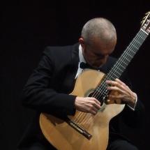 O violonista italiano Fabio Montomoli toca violão clássico em um palco de fundo escuro. Vestindo terno preto, camisa branca e gravata borboleta, mantém a cabeça inclinada em direção ao instrumento enquanto as duas mãos executam a música sobre as cordas e o braço do violão. A iluminação destaca o músico e o instrumento, reforçando a atmosfera intimista da apresentação.