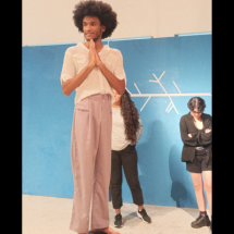 Em uma sala de ensaio, uma pessoa negra de cabelos crespos em estilo black power está em primeiro plano, de pé, com as mãos unidas à frente do peito e o olhar voltado para a esquerda. Veste camiseta branca e calça lilás. Ao fundo, diante de um painel azul com um diagrama de causa e efeito desenhado em branco, outras duas pessoas observam a atividade.