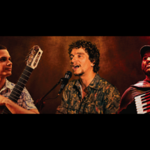 Os integrantes do trio Três em Travessia aparecem lado a lado sobre um fundo em tons de marrom e laranja. À esquerda, o violonista de sete cordas Igor Souza segura o instrumento e olha para o centro. Ao centro, o cantor e pianista Gabriel Ruiz canta ao microfone. À direita, o sanfoneiro e cantor Rafael Meninão toca sanfona enquanto olha para cima com um leve sorriso.