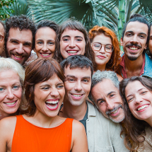 Integrantes do grupo vocal Vozerê sorriem reunidos para uma fotografia em close. O grupo é formado por doze pessoas de diferentes idades, posicionadas bem próximas umas das outras diante de um jardim com folhagens ao fundo. As expressões alegres e a variedade de sorrisos, olhares e gestos transmitem um clima de amizade, acolhimento e celebração.