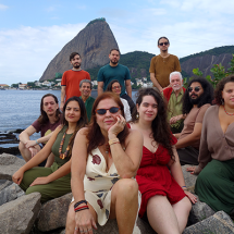 Integrantes do Coral Altivoz posam reunidos sobre pedras à beira da água, tendo ao fundo o Pão de Açúcar e a paisagem da Baía de Guanabara. O grupo é formado por pessoas de diferentes idades, distribuídas em vários níveis, algumas sentadas e outras em pé. Vestindo roupas em tons terrosos, verdes, vermelhos e bege, os integrantes sorriem ou mantêm expressões serenas, compondo um retrato coletivo em ambiente ao ar livre.