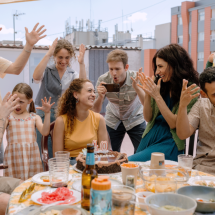 Still de um dos filmes da mostra de cinema espanhol. Um grupo de adultos e uma criança está reunido ao redor de uma mesa durante uma comemoração. Ao centro, uma mulher sorri diante de um bolo com velas que formam o número 40, enquanto as pessoas ao redor levantam as mãos em gestos animados. A mesa está repleta de pratos, copos e bebidas, e a cena acontece em uma área externa cercada por edifícios.