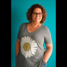 Retrato da escritora Luciana Belém. Ela aparece em pé, sorrindo para a câmera, com cabelos castanhos cacheados e óculos. Veste blusa cinza com a estampa de uma grande margarida branca e mantém as mãos nos bolsos. Ao fundo, uma parede azul-turquesa contrasta com a roupa.
