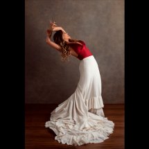 Retrato da bailarina Eliane Carvalho em pose de dança. Ela inclina o corpo para trás, com o rosto voltado para cima e os braços elevados e curvados sobre a cabeça. Veste blusa vermelha e uma longa saia branca com babados, cuja cauda se espalha pelo chão de madeira ao seu redor. Ao fundo, uma parede em tom marrom neutro destaca a figura da bailarina.