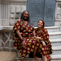 As contadoras de histórias Claudia Marques e Pâmela Cristina sorriem sentadas nos degraus de uma escadaria de pedra diante de um edifício histórico. As duas vestem roupas com a mesma estampa em tons de vinho, amarelo e azul, criando uma identidade visual marcante. O ambiente de arquitetura clássica reforça o clima acolhedor da fotografia.