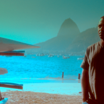 Still do filme Racismo. Um homem aparece em primeiro plano, à direita, usando camiseta escura e olhando para fora do quadro. Ao fundo, vê-se a Baía de Guanabara, com o Pão de Açúcar em destaque entre os morros. À esquerda, parte de uma estrutura com equipamentos náuticos ocupa a lateral da imagem. A cena tem predominância de tons azulados e esverdeados.