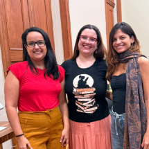 As escritoras e mediadoras Beatriz Ras, Carolina Pessoa e Nicole Ayres posam juntas, sorrindo para a câmera, em uma sala do CCJF. As três aparecem lado a lado, em pé, diante de portas de madeira, vestindo roupas em tons de vermelho, preto, rosa e cinza. Ao fundo, mesas e cadeiras compõem o ambiente.