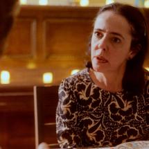 Still do filme O que resta de nós?. A atriz Sílvia Buarque aparece sentada à mesa, voltada para uma pessoa desfocada em primeiro plano. Com expressão atenta e séria, olha diretamente para o interlocutor enquanto parece conversar. Veste uma blusa estampada em preto e branco. Ao fundo, um ambiente iluminado por pequenas luzes em tons quentes reforça a atmosfera intimista da cena.
