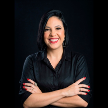 Retrato da professora Tati Vidal. Ela aparece sorrindo para a câmera, com os braços cruzados, cabelos escuros curtos e veste camisa preta. O fundo também é preto, destacando o rosto e as mãos, com unhas pintadas de vermelho.