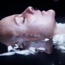 Still de curta-metragem da Mostra Vem Ver. Em primeiro plano, uma mulher aparece deitada de olhos fechados, com parte do rosto e dos cabelos mergulhada na água. Flores brancas flutuam ao redor de sua cabeça, e seu rosto se reflete na superfície escura da água.