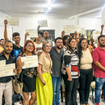 Imagem de referência da Sessão Baixada. Um grande grupo posa reunido em um espaço interno; várias pessoas exibem certificados diante da câmera, enquanto outras aparecem ao centro e ao fundo.
