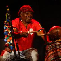 Músico sentado no palco sorri enquanto toca um instrumento de percussão com uma baqueta. Ele veste camisa vermelha e gorro estampado. Ao seu redor, aparecem instrumentos e objetos decorados com tecidos de estampas coloridas, além de um suporte ornamentado com pequenas peças multicoloridas.