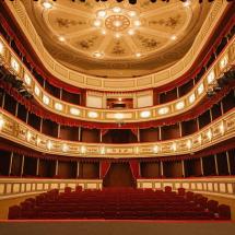 A imagem mostra o interior de um teatro clássico, visto do palco em direção à plateia. O espaço tem assentos estofados em vermelho, três andares de camarotes com detalhes dourados e cortinas vermelhas, além de um teto ornamentado com afrescos e um grande lustre central. A iluminação quente destaca a decoração elegante e simétrica do ambiente.