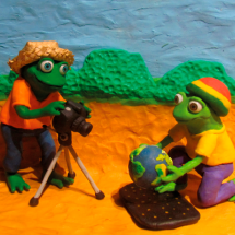 Cena com dois sapos humanoides em estilo de desenho animado, produzidos em massa de modelar. Em um cenário ao ar livre, um deles está diante de uma câmera sobre tripé, enquanto o outro, ajoelhado, segura um pequeno globo terrestre. Ao fundo, aparecem colinas verdes sob um céu azul.