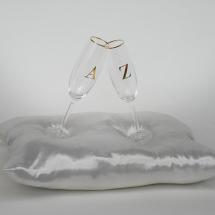 Obra da exposição As Palavras. Duas taças transparentes, com as letras douradas “A” e “Z”, estão inclinadas uma contra a outra, tocando-se pelas bordas. As taças repousam sobre uma almofada branca de tecido acetinado, diante de um fundo claro.