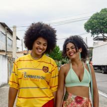Still de Ficção Suburbana. Duas jovens caminham lado a lado por uma rua de bairro, sorrindo. Uma delas veste camisa amarela de futebol e a outra usa top verde-claro e saia florida. Ao fundo, aparecem casas, muros, árvores, postes, fiação e caminhões estacionados ao longo da rua, sob céu nublado.