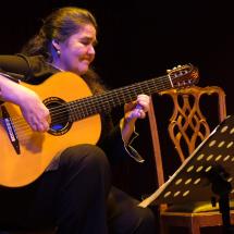 A violonista Mara Lúcia Ribeiro toca sentada no palco, diante de uma estante com partitura. Vestida de preto, ela segura o violão enquanto executa a música, sob iluminação cênica em tons quentes e azulados. Ao lado, há uma cadeira de madeira.