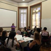 Grupo participa de atividade em uma sala, sentado ao redor de mesas dispostas em conjunto. À frente, uma mulher em pé fala aos participantes, que estão voltados em sua direção. O ambiente tem grandes janelas, tela de projeção e quadro branco, e sobre as mesas aparecem livros, cadernos, garrafas e outros materiais.