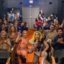Em uma sala de cinema, duas artistas trans posam junto ao público entre as fileiras de poltronas. Uma delas, de vestido rosa, acena para a câmera; a outra, com cabelos amarelos, segura um microfone. Ao redor, pessoas sorriem, aplaudem e acenam para a foto.