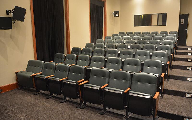 A foto mostra um cinema pela visão de quem está no tablado. Há cadeiras pretas com detalhes em madeira clara. O chão é cinza, de carpete, e há uma escada na parte direita da imagem. É possível ver janelas com longas cortinas pretas. O ambiente é bem iluminado com luz artificial.