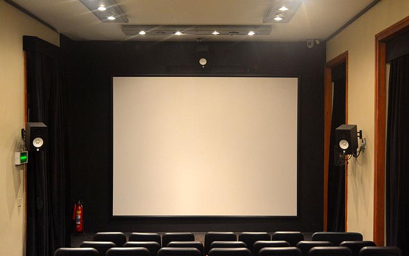 A foto mostra um cinema pela visão do público. Há cadeiras pretas com detalhes em madeira. É possível ver duas caixas de som e uma tela branca. As paredes são beges e há cortinas pretas nas janelas. O ambiente é bem iluminado. É o cinema do Centro Cultural Justiça Federal.