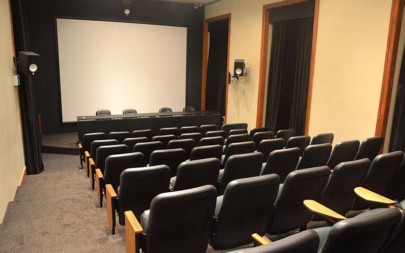 A foto mostra um auditório pela visão do público, com um telão na frente. Há cadeiras pretas com detalhes em madeira, uma mesa central em cima do tablado, também com cadeiras. As paredes são beges e há cortinas pretas nas janelas. O ambiente é bem iluminado. É o cinema do Centro Cultural Justiça Federal.