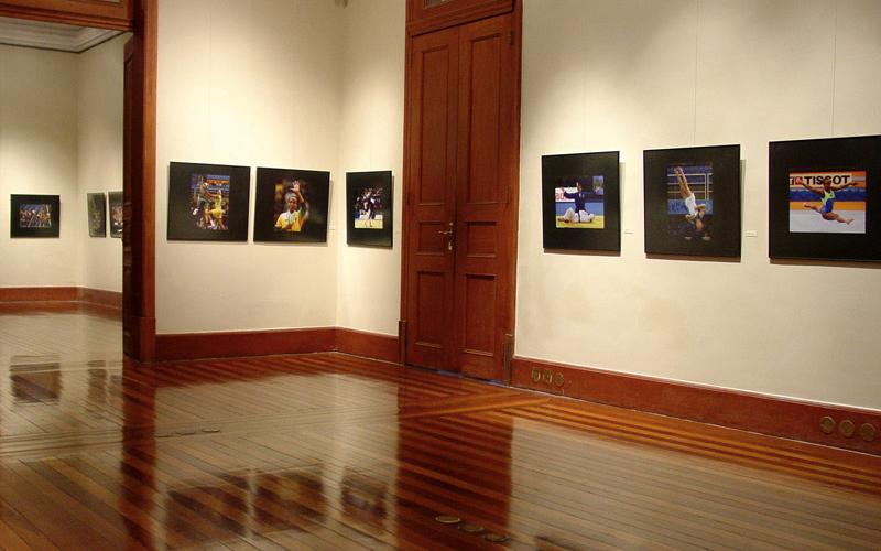 Na foto, está em destaque uma porta alta de madeira escura. É possível ver alguns quadros de atletas nas paredes e uma outra porta aberta do lado esquerdo. O chão, também de madeira, é do mesmo tom das portas. Ela retrata parte das galerias do Centro Cultural Justiça Federal.