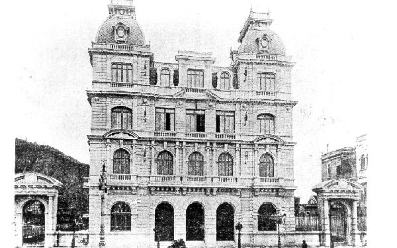 Fotografia, em preto e branco do prédio que hoje abriga o Centro Cultural Justiça Federal. A fotografia está bem pixelizada como se tratasse de uma fotografia antiga que foi digitalizada. A arquitetura do prédio é eclética, composta por 4 andares, portas e janelas arredondadas e duas torres nas extremidades direita e esquerda do último andar. Em cada lado do prédio, há um pórtico com o mesmo estilo arquitetônico.