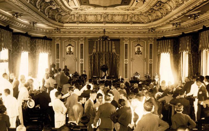Fotografia em sépia de um evento com diversas pessoas de pé e vestidas com traje de época. A sala tem um estilo colonial, o teto e a parede de fundo possuem detalhes clássicos, as paredes laterais possuem 3 janelas cada, com cortinas abertas por onde passam bastante luz.