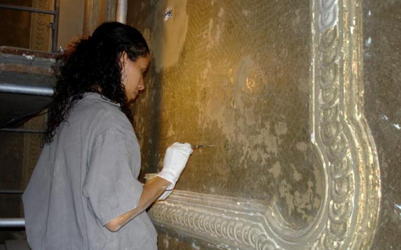 Na imagem, uma mulher negra com cabelos pretos enrolados, blusa cinza e um par de luvas aparece restaurando uma parede antiga.