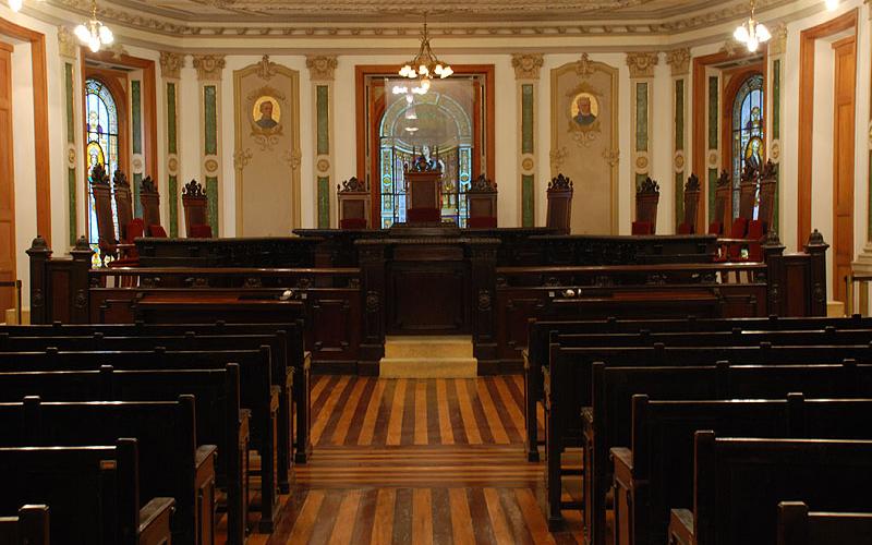 A foto mostra um recorte de uma sala da época colonial, com cadeiras para o público e um parlamento, tudo em madeira. Há lustres dourados; as paredes e teto são desenhadas e com arabescos clássicos. O local é a Sala de Sessões do Centro Cultural Justiça Federal.