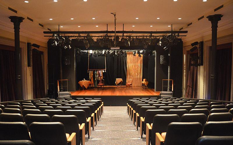 A foto mostra um teatro pela visão do público. No centro da tela há o palco, onde estão objetos cenográficos e nos cantos direito e esquerdo há cadeiras pretas com detalhes em marrom. Há uma escadaria preta indo ao encontro do palco. O teatro é do Centro Cultural Justiça Federal.