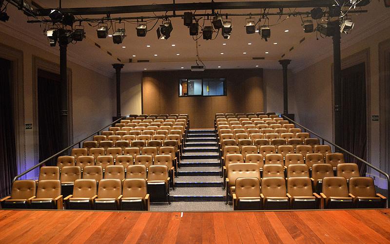 A foto mostra um teatro pela visão de quem está no palco (de madeira) se apresentando. No centro, há uma escadaria cinza e nos cantos direito e esquerdo há cadeiras com detalhes em marrom, enfileiradas. No teto é possível ver luzes características para peças teatrais. O teatro é do Centro Cultural Justiça Federal. 