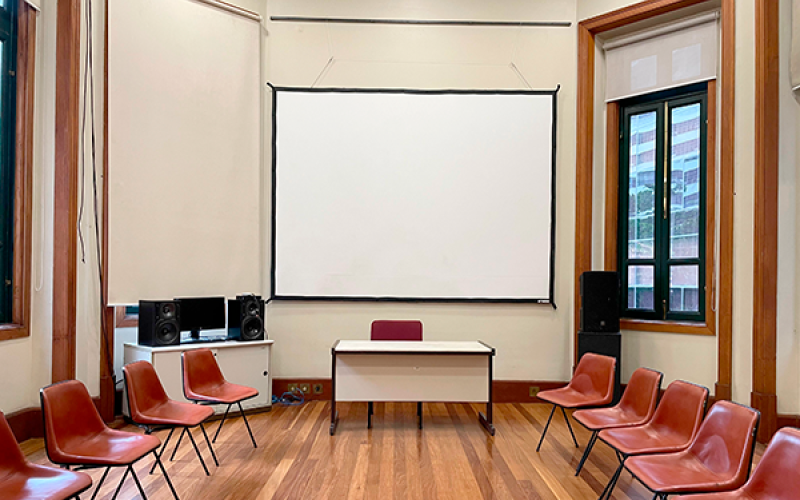 A imagem mostra uma sala retangular com piso de madeira e paredes claras. No centro, há uma mesa branca com uma cadeira vermelha em frente a uma grande tela de projeção. Cadeiras de plástico vermelho estão dispostas em semicírculo ao redor, sugerindo um ambiente de apresentação ou reunião. À esquerda, um móvel branco com caixas de som, e à direita, uma janela que permite entrada de luz natural. A sala parece organizada e adequada para palestras ou discussões em grupo.