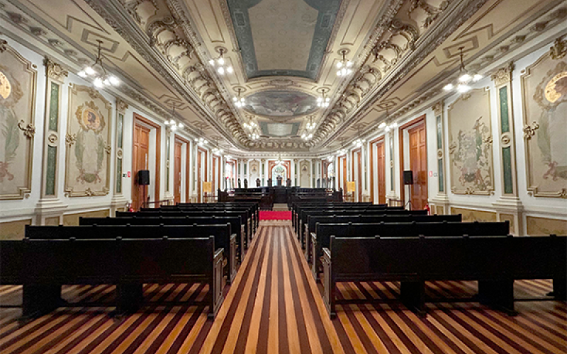 Imagem da Sala de Sessões do Centro Cultural Justiça Federal (CCJF), com vista frontal para o espaço. A foto mostra fileiras de bancos de madeira escura alinhadas em direção ao palco ao fundo, onde se vê uma mesa cerimonial e um vitral colorido. O piso tem listras em tons claros e escuros, o teto é ricamente decorado com afrescos e molduras em relevo, e as paredes exibem painéis artísticos com detalhes dourados. A iluminação clara ressalta a elegância e imponência do ambiente.