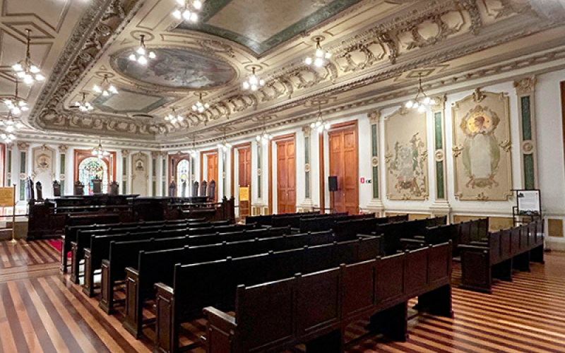 Vista lateral da Sala de Sessões do Centro Cultural Justiça Federal (CCJF), com fileiras de bancos de madeira escura sobre um piso listrado em tons claros e escuros. As paredes são decoradas com grandes painéis artísticos e detalhes dourados. O teto é ricamente adornado com molduras e afrescos, e o espaço é iluminado por elegantes lustres. A sala transmite sofisticação e valor histórico.