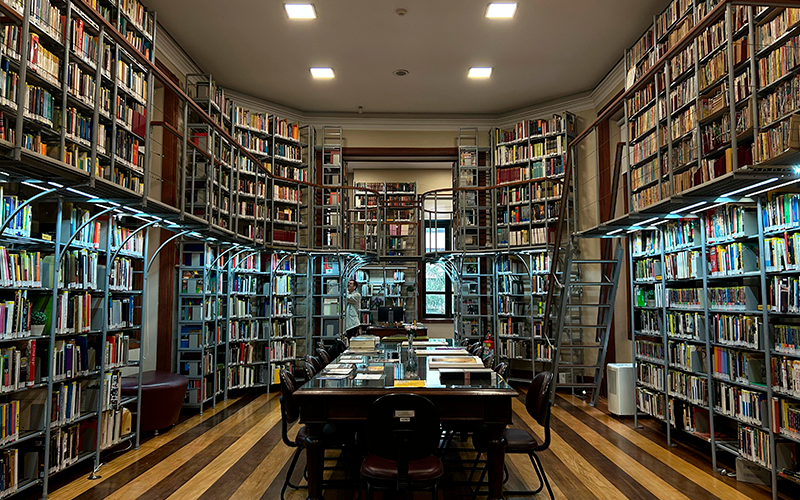 Vista frontal da biblioteca do Centro Cultural Justiça Federal (CCJF). A imagem mostra o ambiente amplo e simétrico, com estantes metálicas repletas de livros cobrindo todas as paredes laterais e o fundo da sala até o teto. Ao centro, uma grande mesa retangular de madeira escura está cercada por cadeiras e coberta por livros abertos e empilhados. O piso de madeira listrado e a iluminação clara completam o ambiente silencioso e acolhedor.