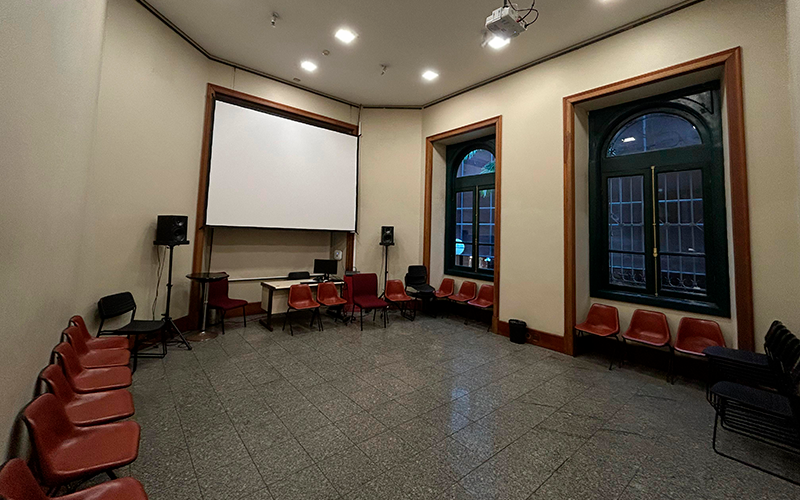 Vista lateral direita da sala de cursos do Centro Cultural Justiça Federal (CCJF), com paredes claras e janelas altas de moldura de madeira escura. Cadeiras plásticas estão dispostas em círculo ao longo das paredes. Ao fundo, há uma tela de projeção, duas caixas de som e uma mesa com equipamentos de áudio e vídeo. O piso é de granito, e o ambiente tem iluminação embutida no teto. A sala está vazia e pronta para receber os alunos.