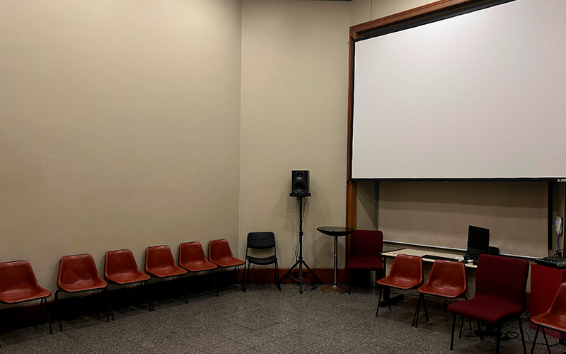Vista lateral esquerda da sala de cursos do Centro Cultural Justiça Federal (CCJF). À direita, uma grande tela de projeção branca domina a parede, acima de uma mesa com equipamentos de áudio e vídeo. Cadeiras plásticas vermelhas estão dispostas em semicírculo ao longo das paredes claras. Uma caixa de som em pedestal e uma pequena mesa redonda completam o ambiente, que tem piso de granito e iluminação embutida. A sala está vazia.
