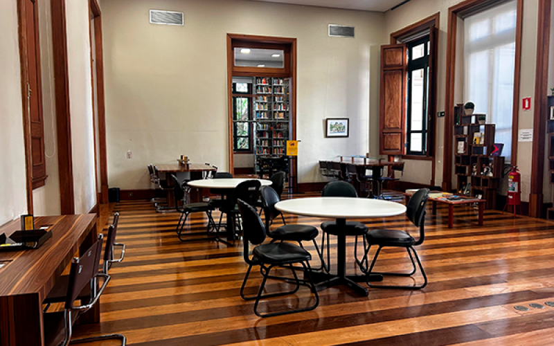 Vista da sala de leitura do Centro Cultural Justiça Federal, com ambiente amplo e iluminado. O espaço tem piso de madeira e paredes claras com detalhes em madeira. Mesas redondas e cadeiras estão organizadas pelo salão, algumas com pessoas sentadas estudando ou lendo. Ao fundo, há estantes e portas altas de madeira, além de janelas que permitem a entrada de luz natural, criando um clima tranquilo e acolhedor.