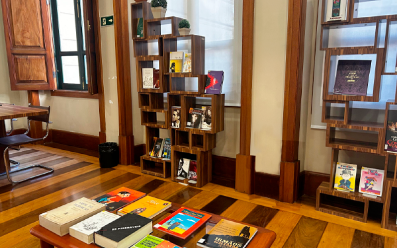 Sala de leitura do CCJF com estantes expositivas de madeira em formato modular, onde livros são exibidos de frente. Em primeiro plano, uma mesa apresenta obras organizadas para consulta. O espaço tem piso de madeira, paredes claras e janelas altas com venezianas, criando um ambiente acolhedor e voltado à leitura.