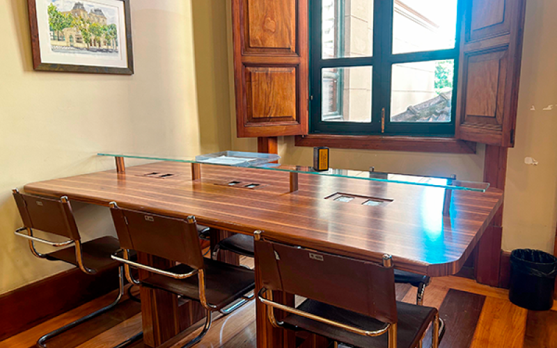 Detalhe da sala de leitura do Centro Cultural Justiça Federal, com mesa retangular de madeira cercada por cadeiras. Sobre a mesa, há alguns materiais organizados. Ao fundo, uma janela ampla deixa entrar luz natural, iluminando o ambiente. O piso de madeira e as paredes claras com molduras reforçam o estilo clássico e acolhedor do espaço, voltado à leitura e estudo.