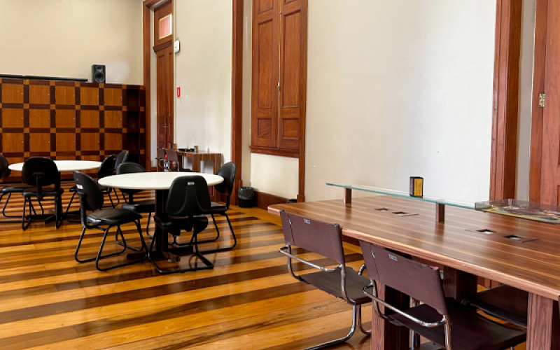 Sala de leitura do CCJF, com mesas redondas e cadeiras distribuídas sobre piso de madeira. À direita, há uma bancada de estudo com divisória de vidro. Ao fundo, veem-se armários de madeira com pequenos compartimentos (escaninhos). O ambiente é interno, organizado e silencioso, voltado à leitura e pesquisa.
