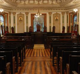 A foto mostra um recorte de uma sala da época colonial, com cadeiras para o público e um parlamento, tudo em madeira. Há lustres dourados; as paredes e teto são desenhadas e com arabescos clássicos. O local é a Sala de Sessões do Centro Cultural Justiça Federal.