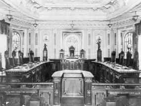 Fotografia em preto em branco de uma sala em estilo colonial. Mobiliário de madeira antigo que faz lembrar uma sala de audiências: ao fundo, mesas e cadeiras de encosto alto e entalhado. À frente e centralizado na foto, há um púlpito de cujos lados se estendem baixas divisórias. Ao fundo de todo esse mobiliário, em uma tonalidade muito clara, quase apagada, teto e paredes com detalhes clássicos.